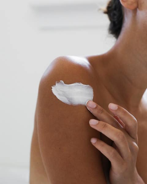 A woman is putting lotion on her shoulder.